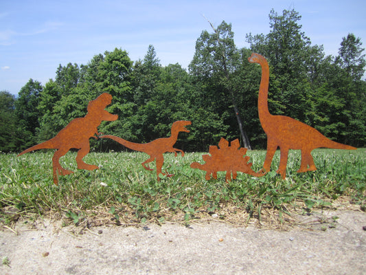 Four rusty metal dinosaur silhouettes in a grassy area. The dinosaurs are a Tyrannosaurus Rex, a Brachiosaurus, a Stegosaurus, and a Velociraptor.  The dinosaurs are all brown and rusty in color and patina over time.