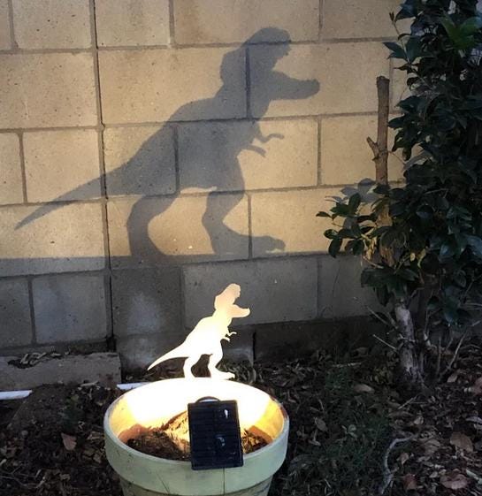A customer example of our metal dinosaur garden stake shown at night with a light in front of it which casts a playful shadow silhouette against the wall behind it.