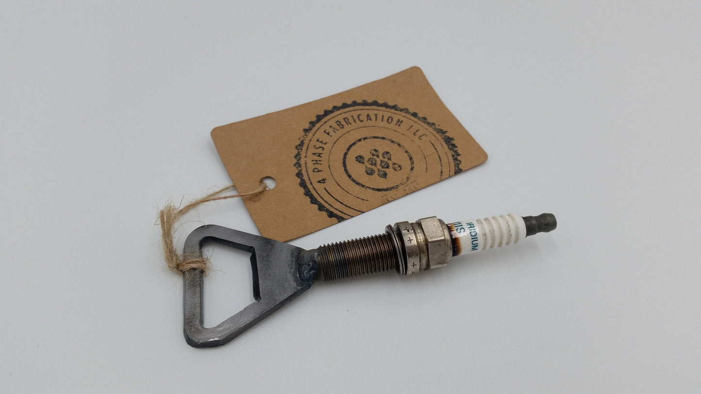 A metal bottle opener with a spark plug attached to it. The bottle opener is silver and has a triangular shape.