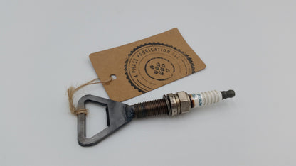 A metal bottle opener with a spark plug attached to it. The bottle opener is silver and has a triangular shape.