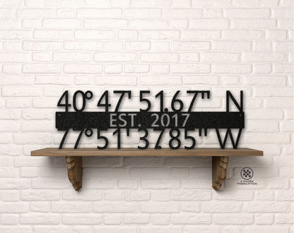 May include: Black metal sign with coordinates 40°47'51.67" N and 77°51'3785" W, EST. 2017. The sign is displayed on a wooden shelf against a white brick wall. The sign is a decorative item.